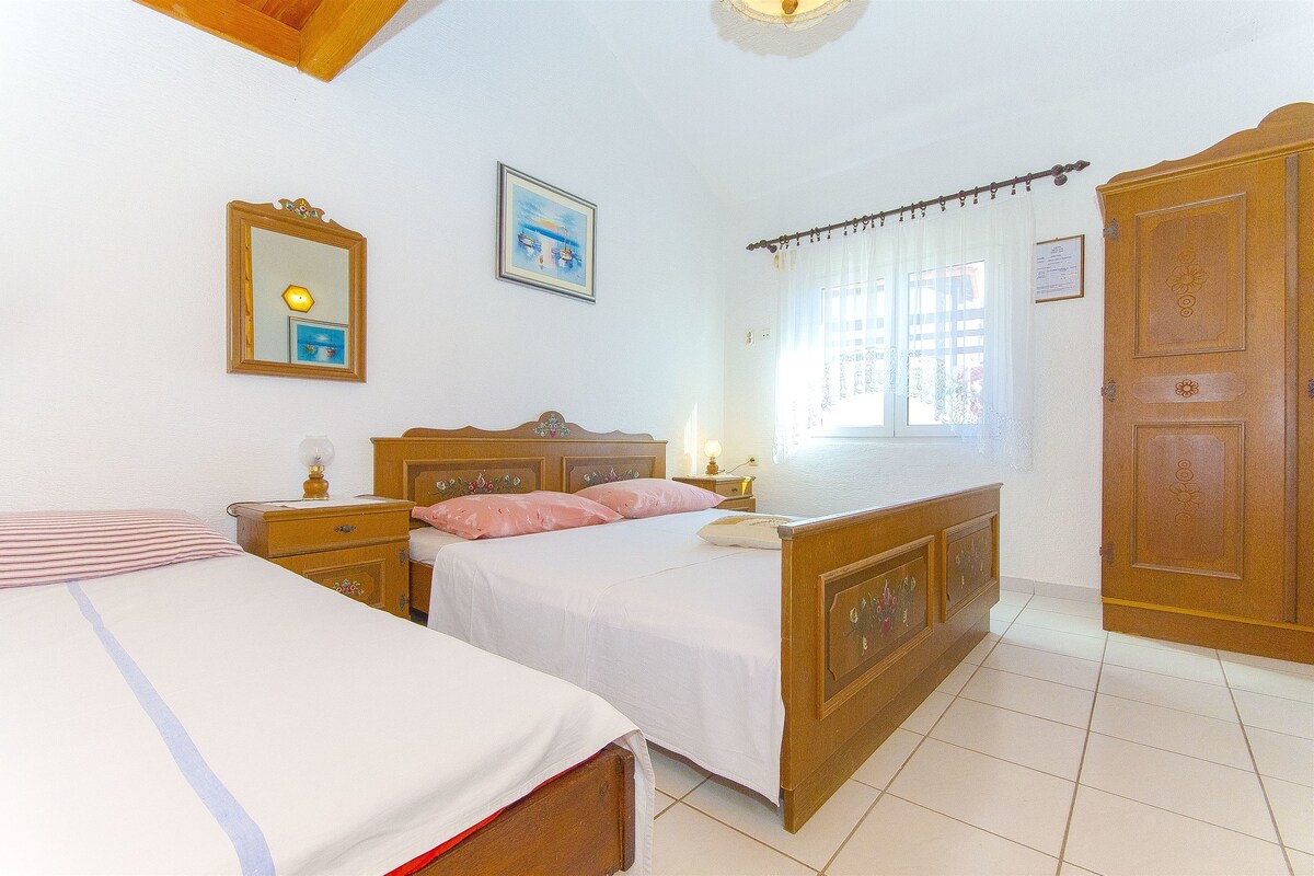 A bedroom with two single beds, wooden headboards, bedside tables with lamps, a window with curtains, and a wooden wardrobe.