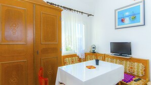 A room with a wooden wardrobe, a white table with a colorful placemat, a bench with floral cushions, a television on a wooden stand, and a framed picture of boats on the wall. - Apartments Pera (Razanj)