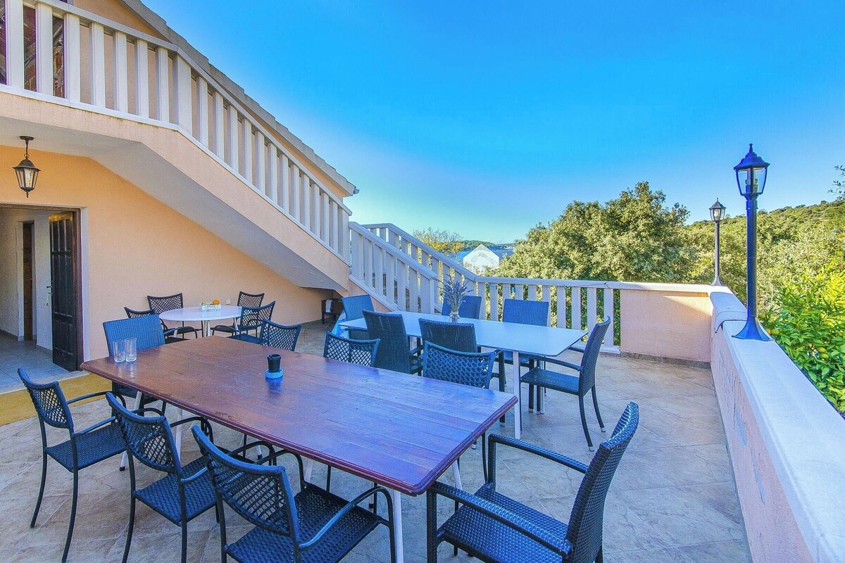 An outdoor patio with a wooden table and chairs, a staircase, and a view of greenery.