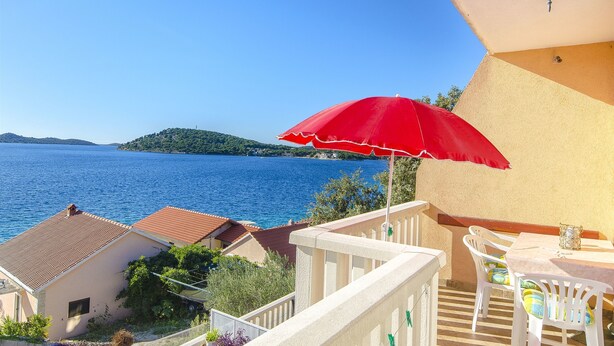 A balcony with a red umbrella, white chairs, and a table overlooking a body of water. - Apartments Pera (Razanj)