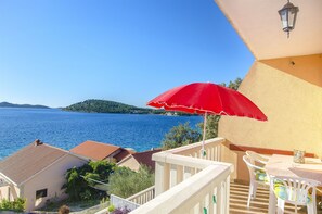 A balcony with a red umbrella, white chairs, and a table overlooking a body of water. - Apartments Pera (Razanj)