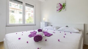 Ein ordentlich gemachtes Bett mit aufgerollten Handtüchern und verstreuten violetten Blumen, ein weißer Nachttisch, eine Wandlampe und ein Fenster mit Blick auf Gebäude.