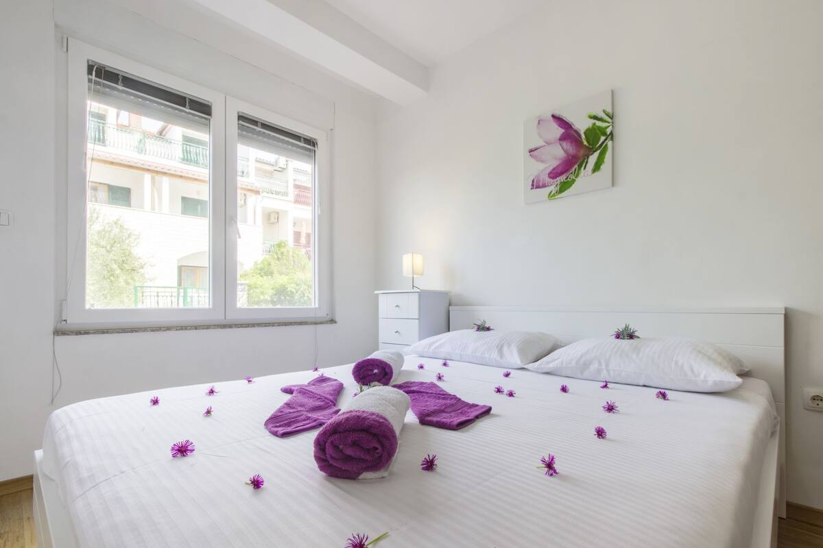 Un lit bien fait, avec des serviettes pliées et des fleurs violettes éparpillées, une table de chevet blanche, une lampe fixée au mur et une fenêtre donnant sur des immeubles.