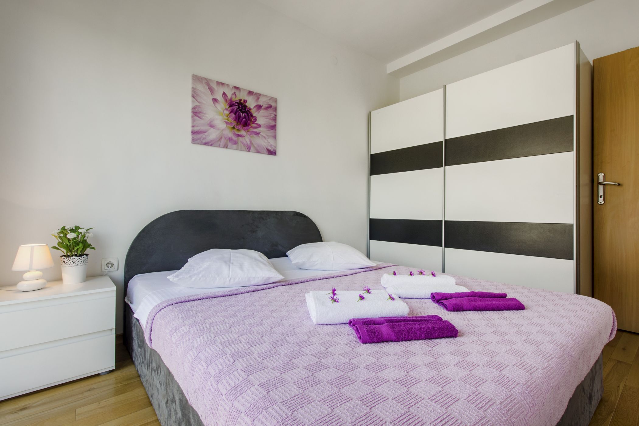 A neatly made bed with a purple blanket, white pillows, and towels, a bedside table with a lamp and a plant, and a large wardrobe with a black stripe.