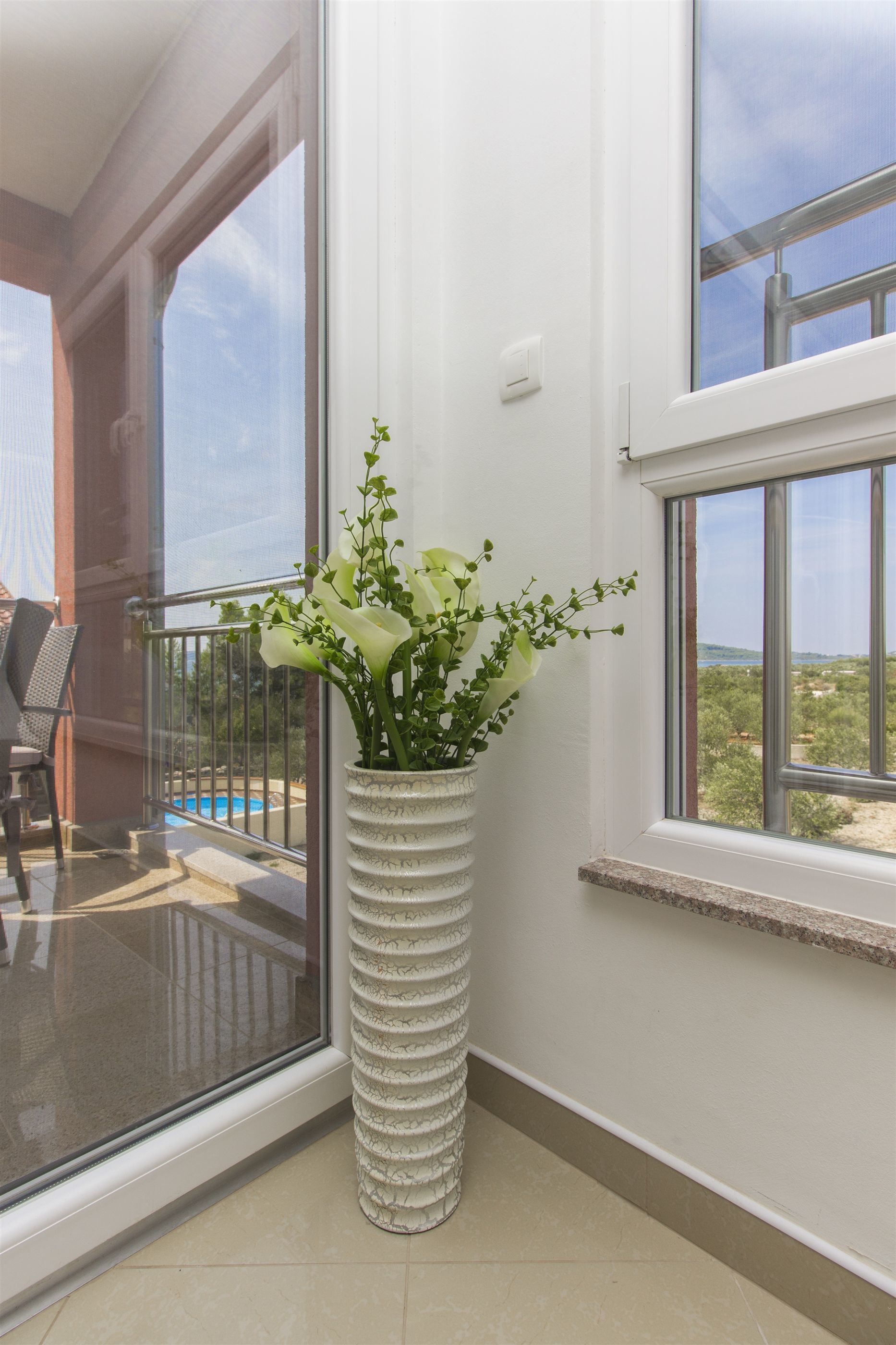 A white vase with flowers on a balcony with a glass door and a view of a pool and greenery.