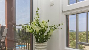 Eine weiße Vase mit Blumen auf einem Balkon mit Glastür und Blick auf Pool und Grünflächen.