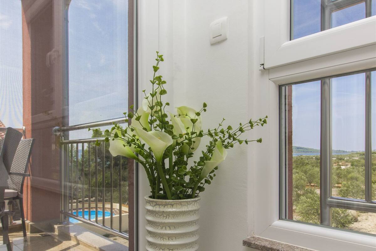 Un vase blanc orné de fleurs, placé sur un balcon doté d’une porte vitrée, d’où l’on peut admirer une piscine et de la verdure.
