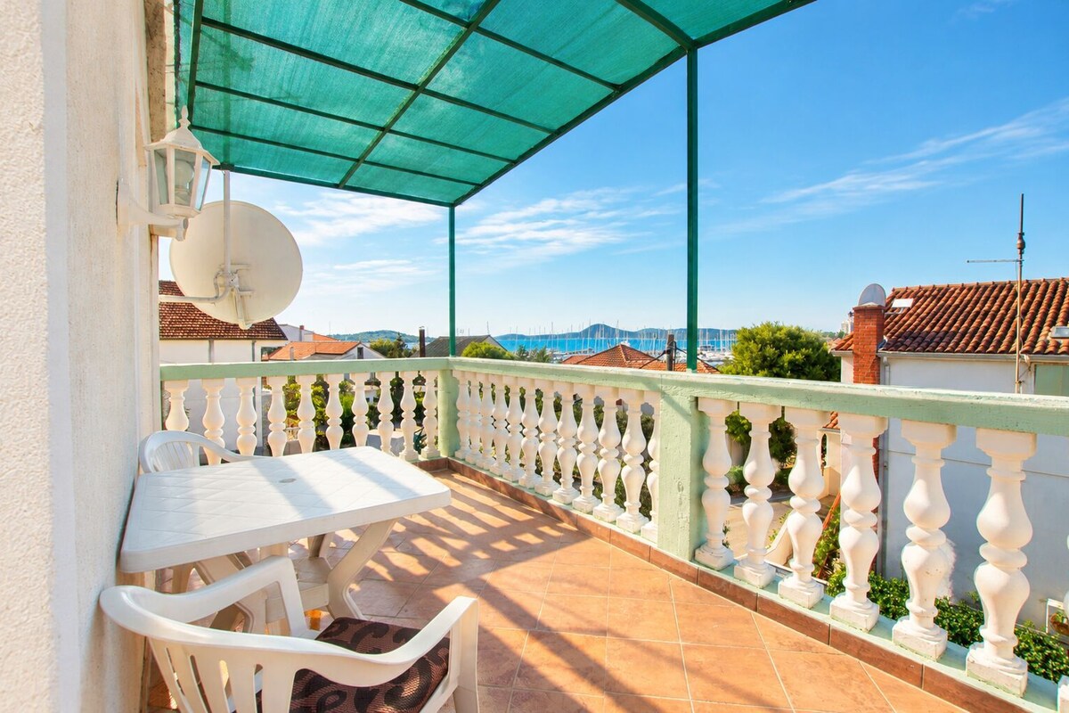 A balcony with a table and chairs, a satellite dish, and a view of the sea and buildings.