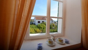 Une fenêtre donnant sur un jardin et des bâtiments, une table blanche avec des tasses et un pichet, et des rideaux orange.