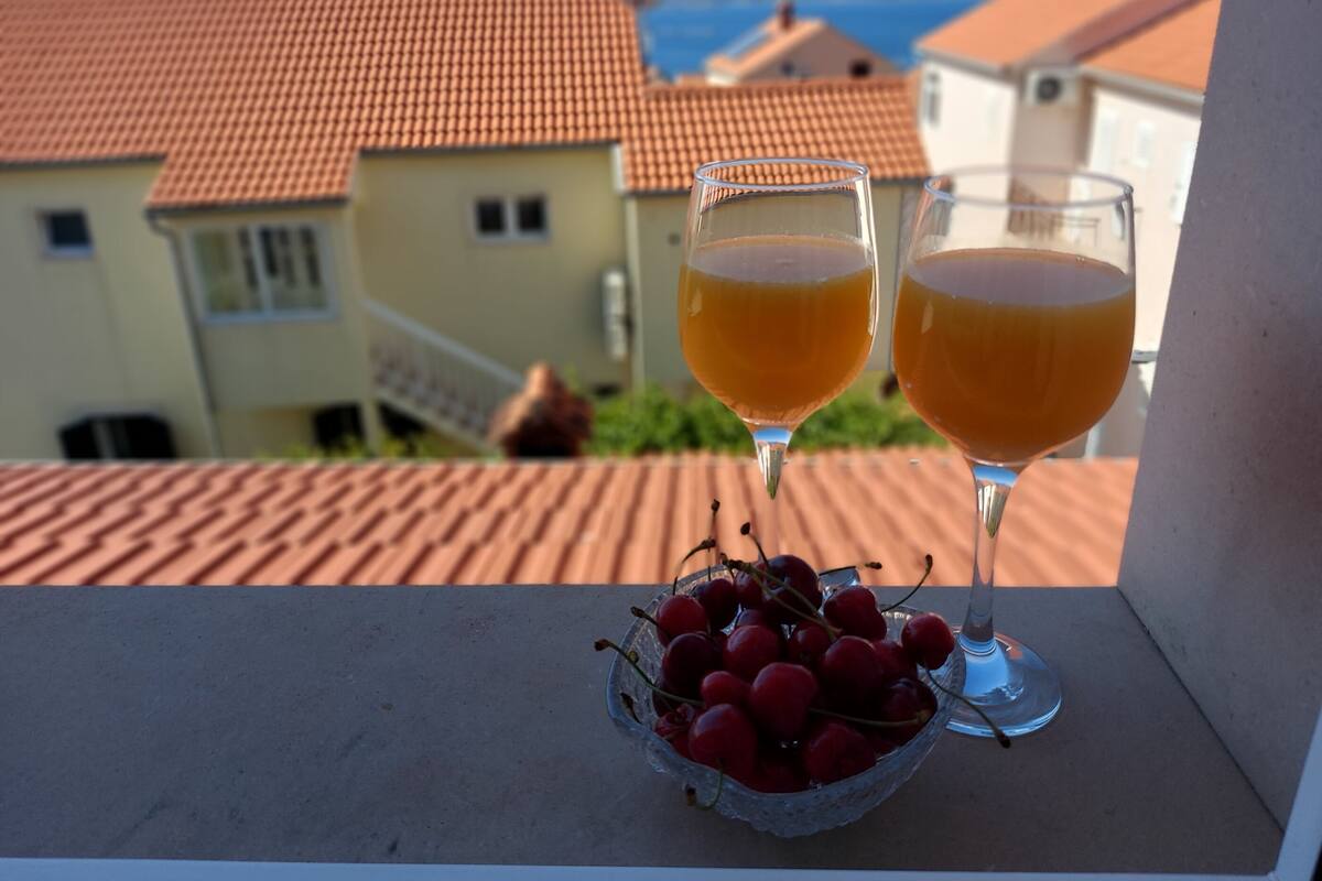 Two glasses of orange juice on a balcony with a bowl of cherries and a view of houses with red roofs.