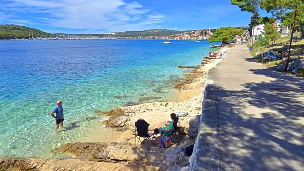 A coastal scene with clear water, a sandy beach, and a road with parked cars.