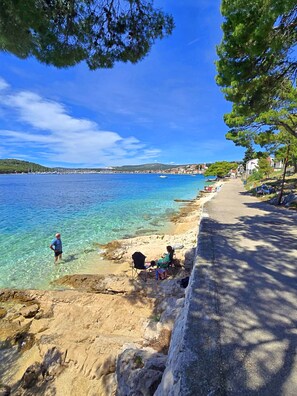 A coastal scene with clear water, a sandy beach, and a road with parked cars. - Apartment Vlatka (Rogoznica)