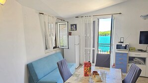 A room with a blue sofa, a dining table with chairs, a TV, and a view of the sea through a balcony.