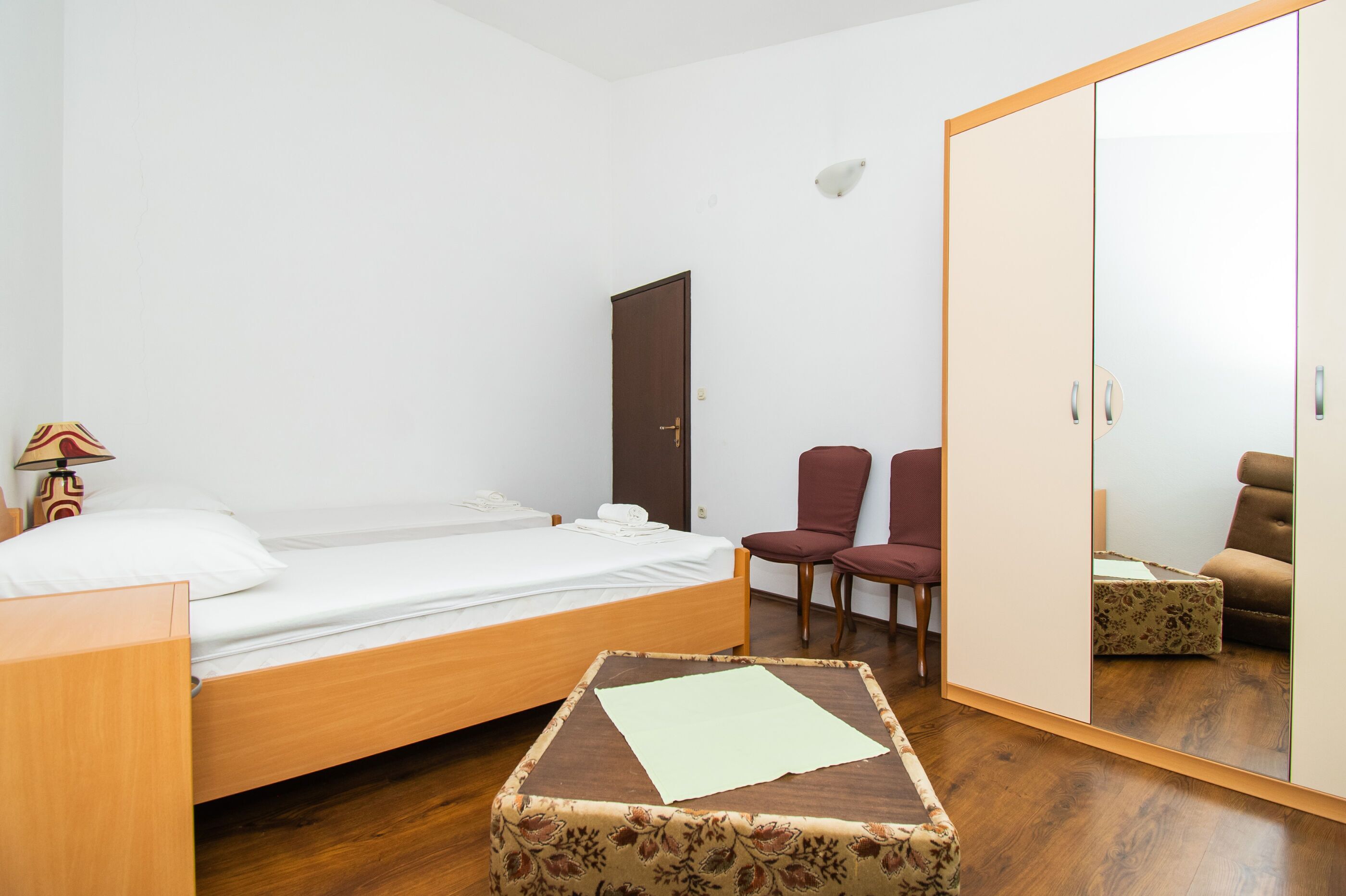 Una camera da letto piccola e ordinata con un letto singolo, un comodino in legno, una sedia, un armadio e uno specchio.