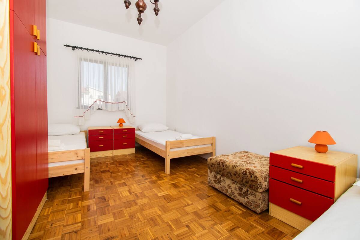 A small, well-lit bedroom with two single beds, a red wardrobe, a wooden nightstand with a lamp, and a patterned ottoman.