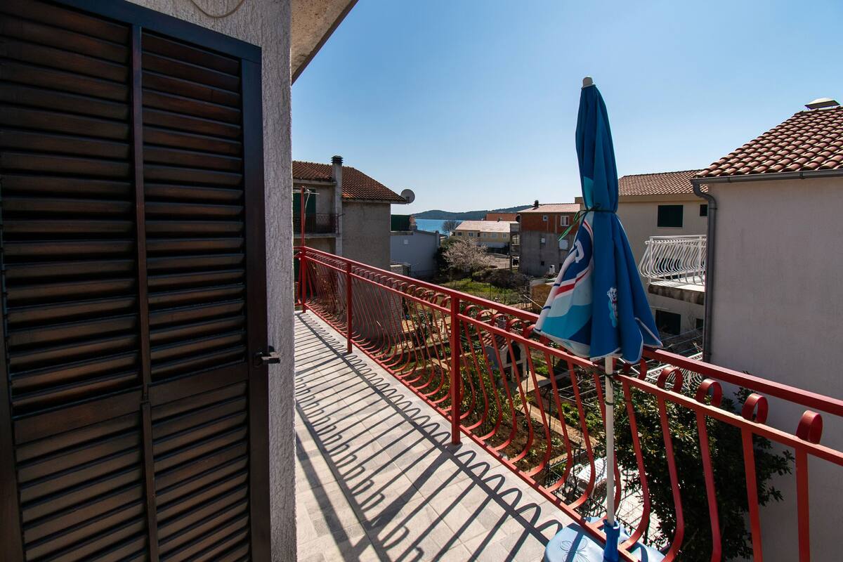 A balcony with a red railing, a blue umbrella, and a view of neighboring buildings.