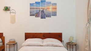 A bedroom with a wooden headboard, a bed with white bedding, two bedside tables with lamps, and a wall-mounted artwork depicting a sunset over a pier. - Apartments Ivana (Nin)