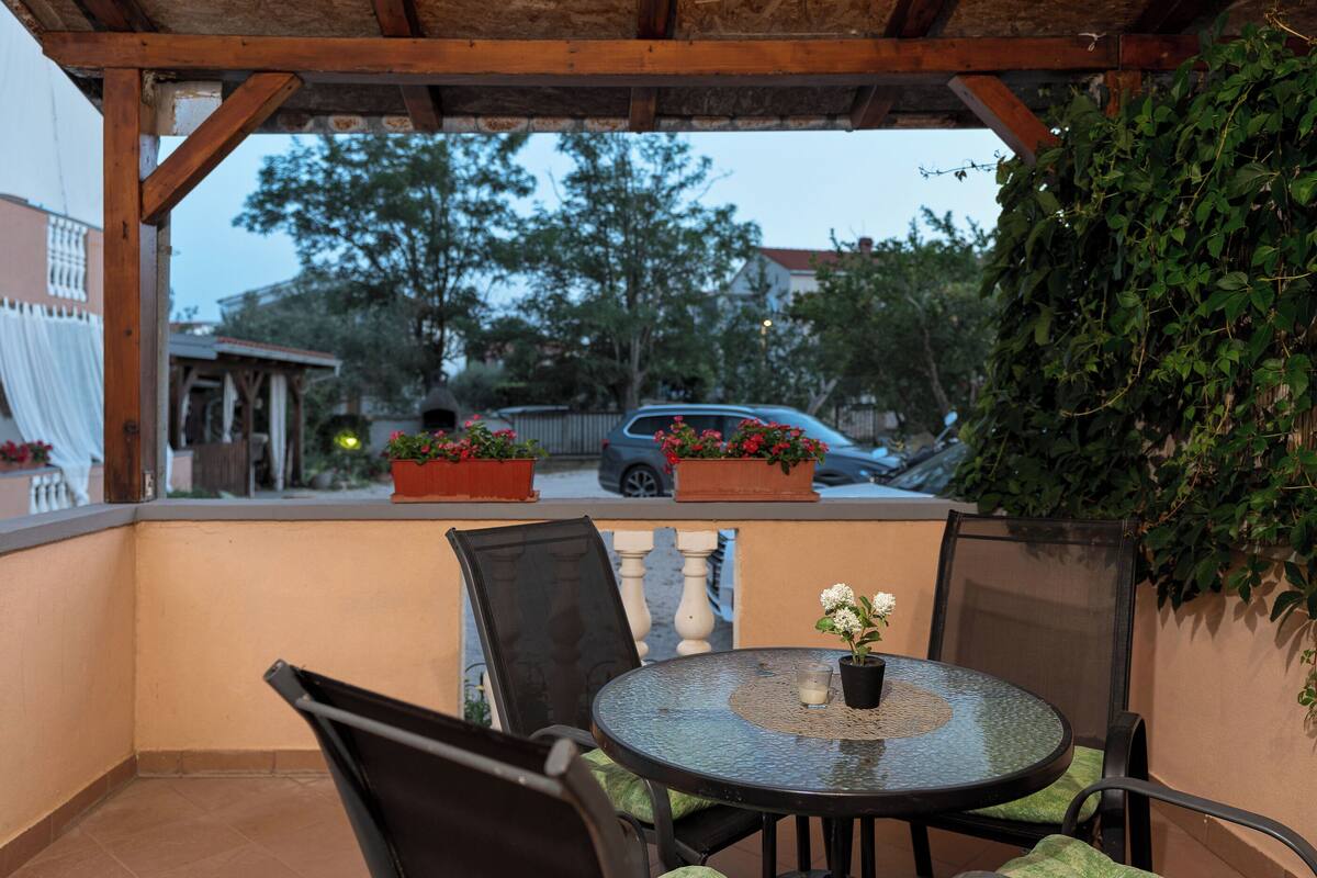 Un portico coperto con un tavolo rotondo e sedie, piante in vaso e vista su una strada con auto e alberi.