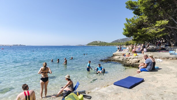 A beach with clear blue water, people swimming and sunbathing, and a rocky shoreline with trees. - Apartments Vlasta (Tribunj)