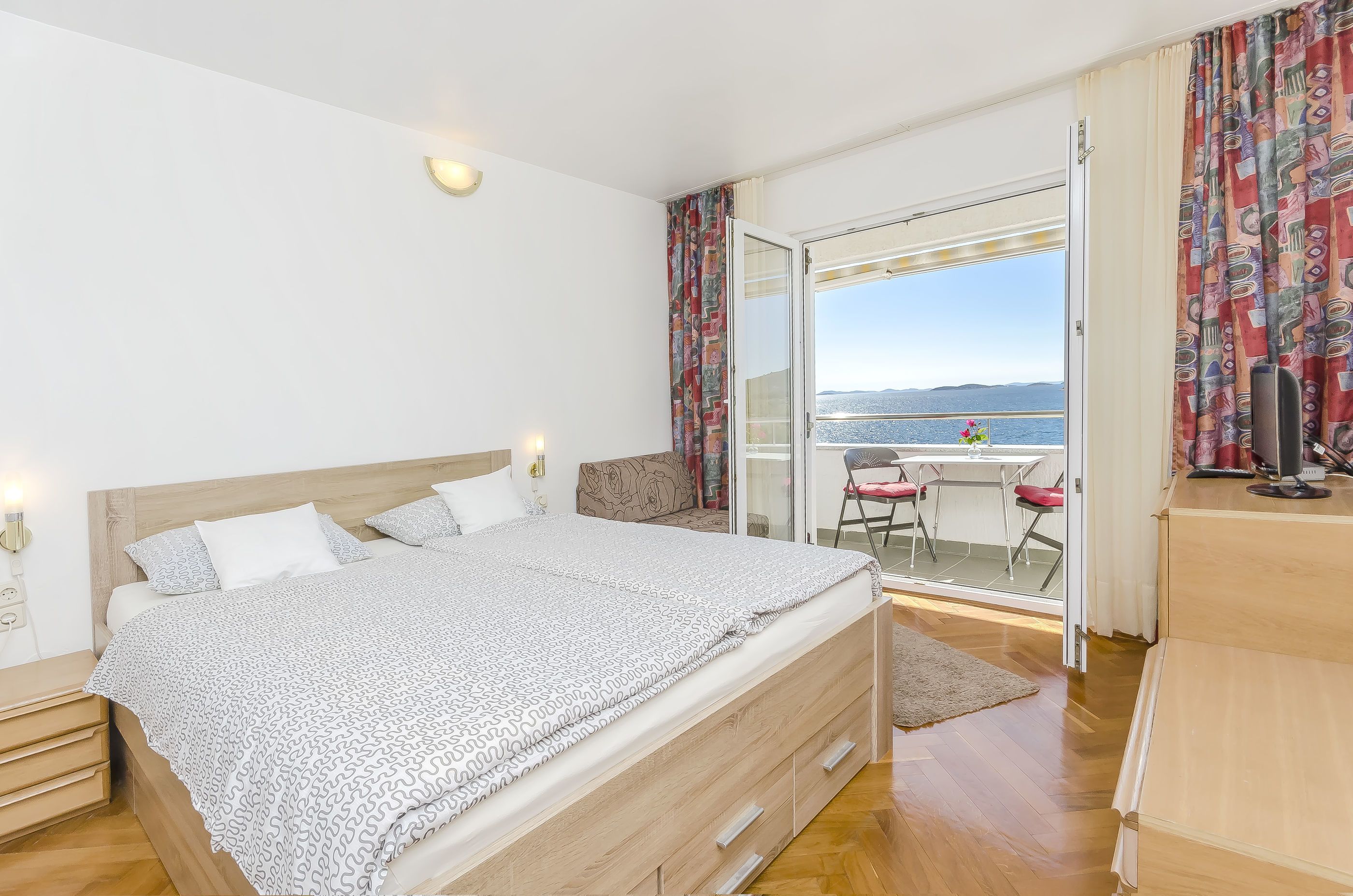 Une chambre à coucher avec un lit, un petit bureau, une chaise et une vue sur la mer, visible à travers une porte vitrée.