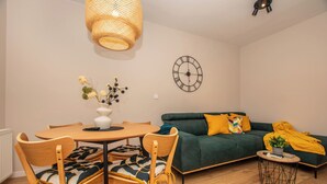 A modern living room with a round wooden dining table, two wooden chairs, a green sofa with yellow cushions, a round side table with a plant, and a wall clock.