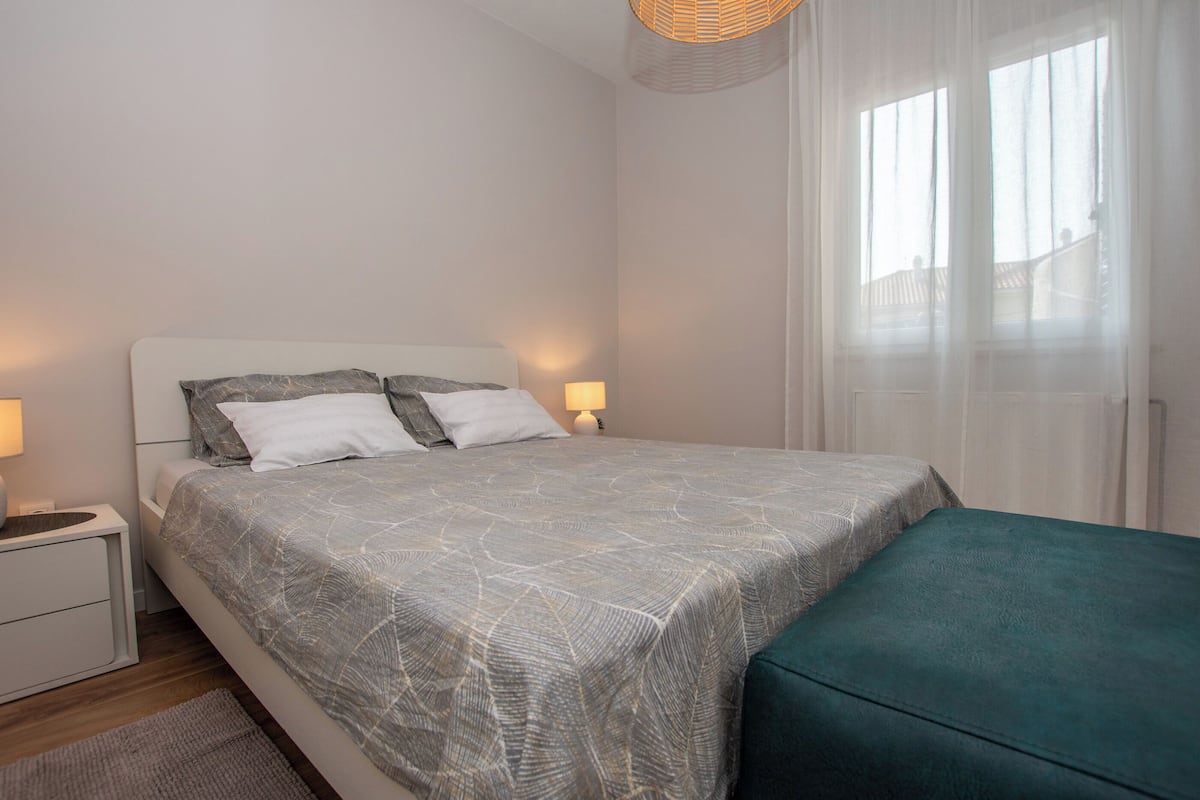 A neatly made bed with a patterned bedspread, a bedside table with a lamp, and a window with sheer curtains.