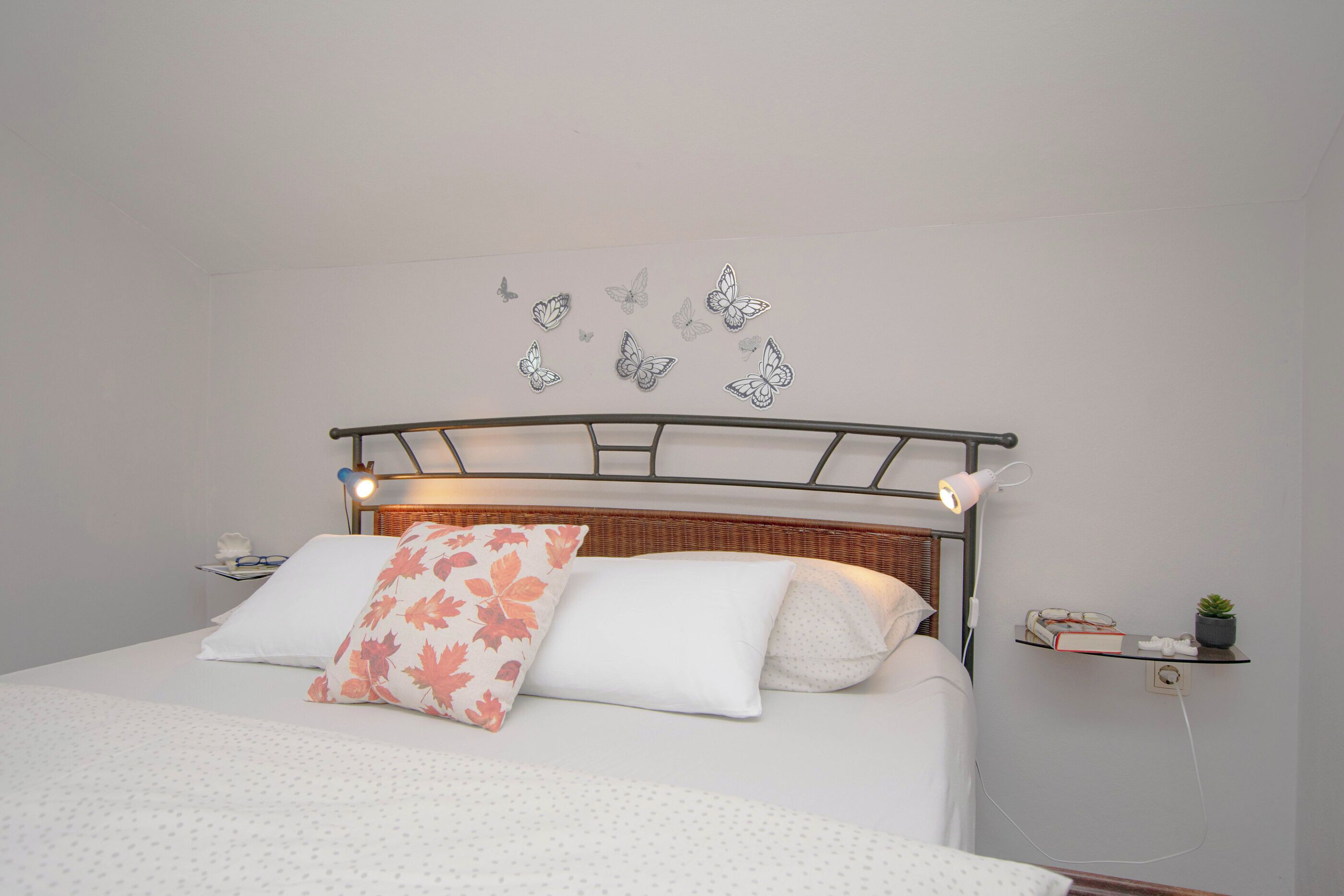 Una cama bien tendida con cabecera metálica, ropa de cama blanca y un cojín decorativo con estampado de hojas.