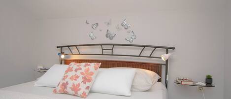 Una cama bien tendida con cabecera metálica, ropa de cama blanca y un cojín decorativo con estampado de hojas.
