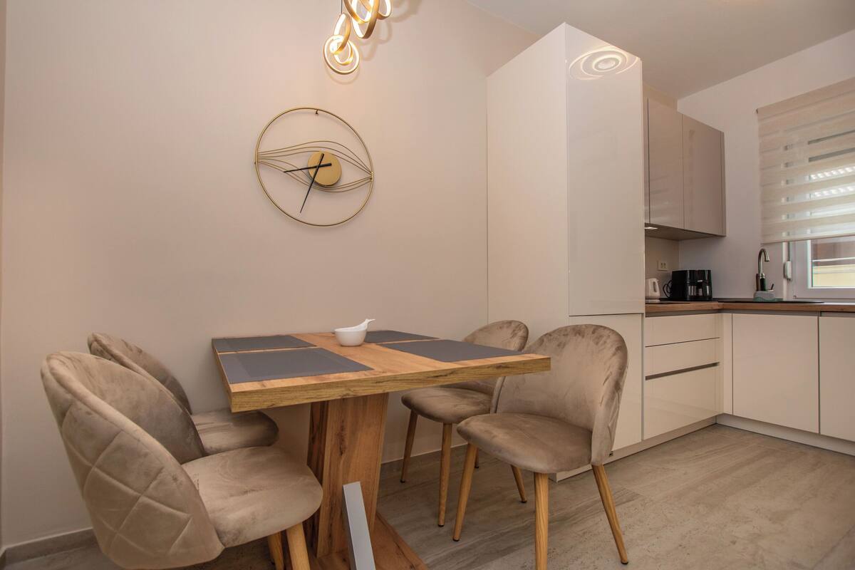 A modern dining area with a wooden table, four cushioned chairs, a wall clock, and a kitchen in the background.
