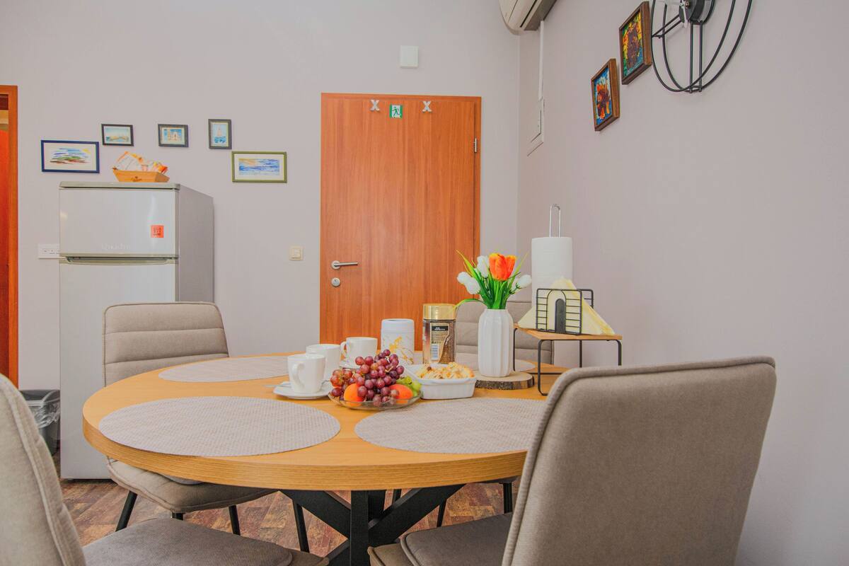 A dining area with a round table, chairs, a refrigerator, a door with a sign, and wall-mounted pictures.
