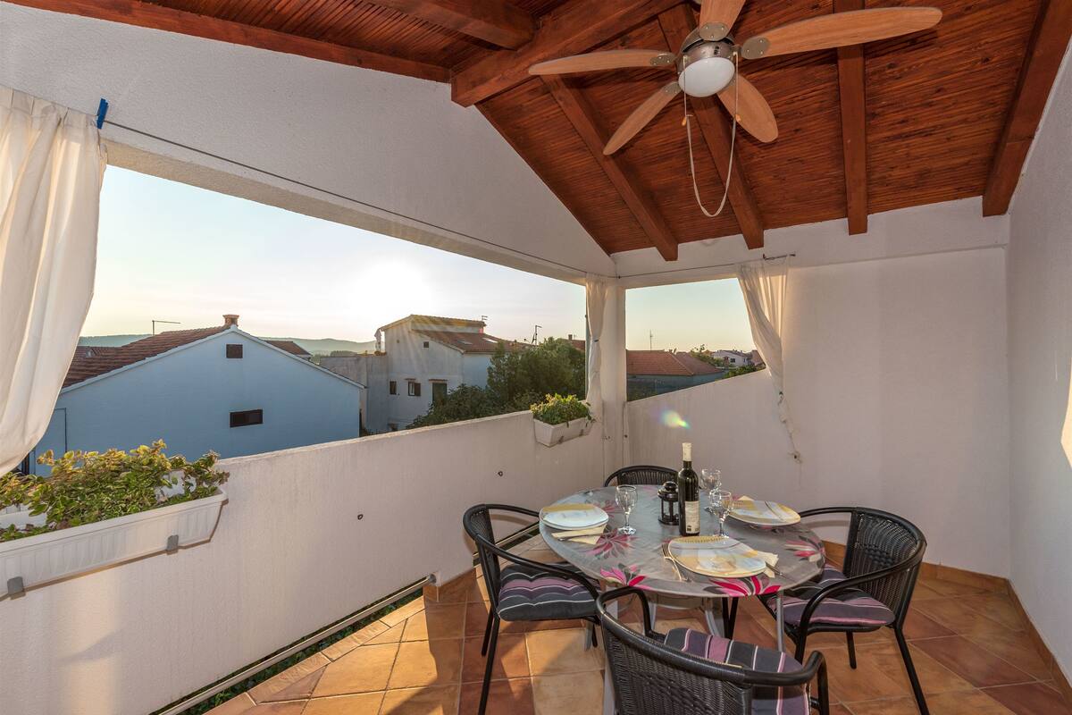 A covered outdoor patio with a table set for dining, a ceiling fan, and a view of the surrounding buildings.