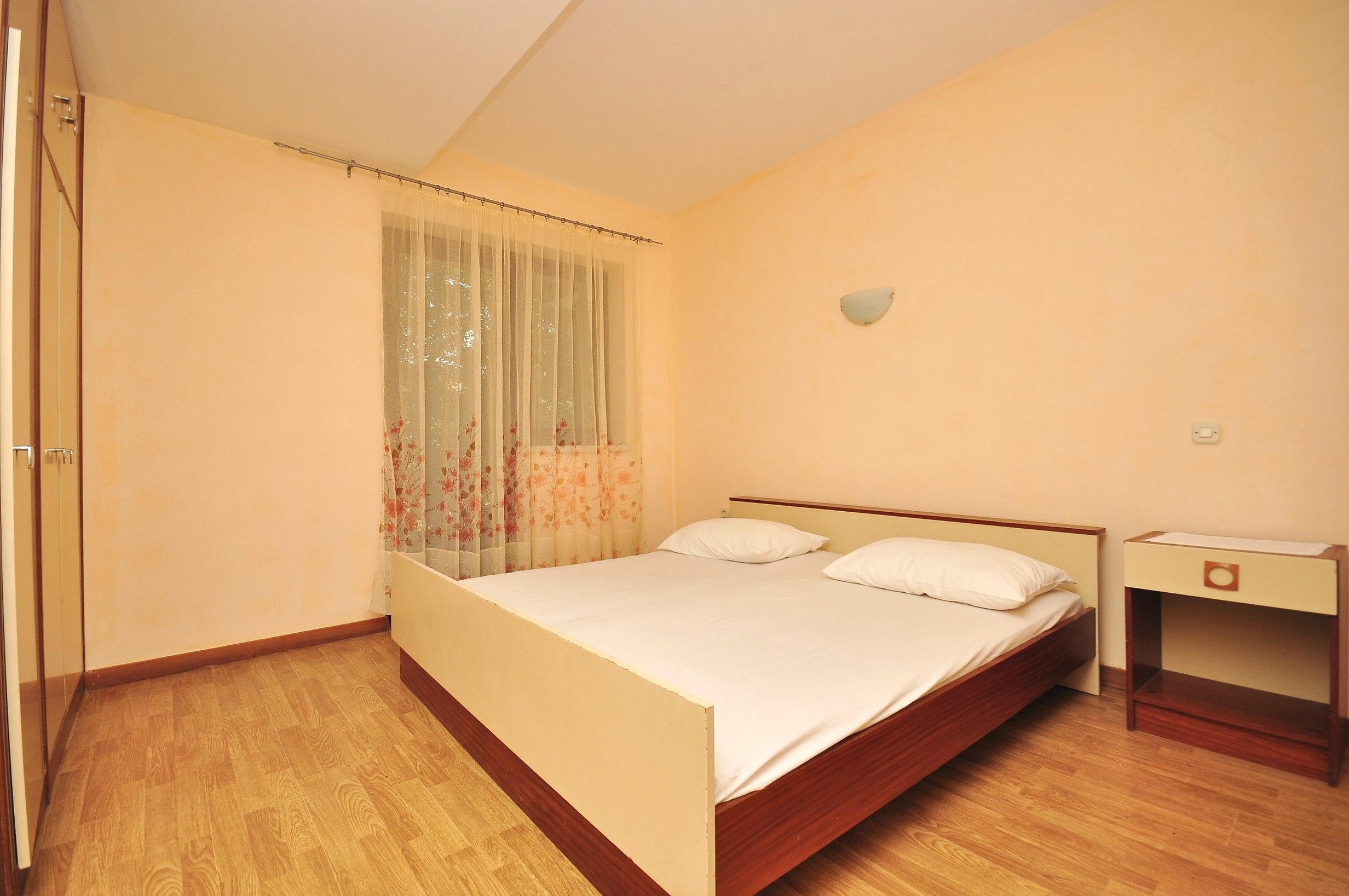 A bedroom with a wooden bed, a nightstand, a window with floral curtains, and a light switch on the wall.