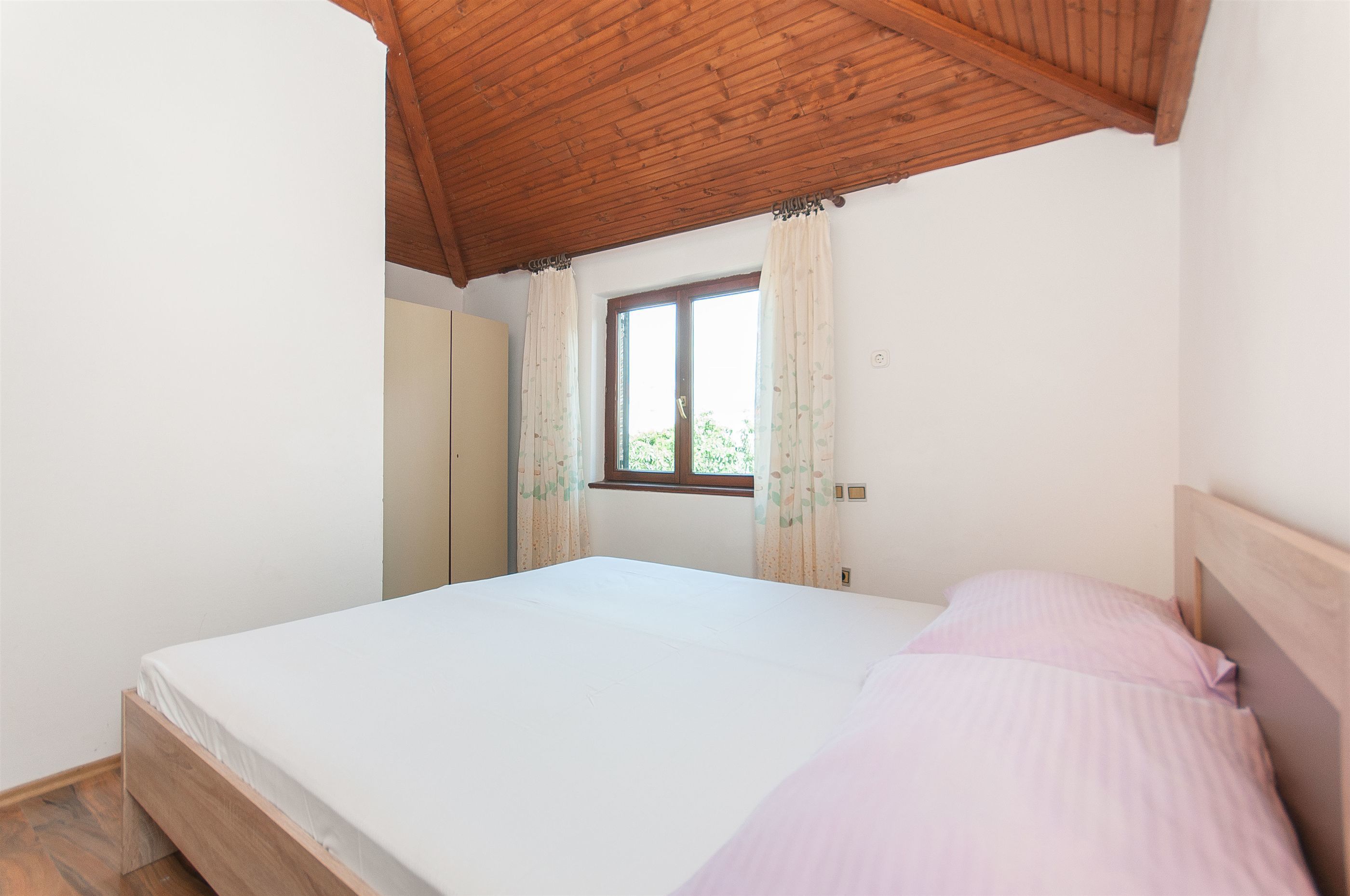 Un dormitorio con techo de madera, una cama individual con una manta rosa, una ventana con cortinas y un armario.