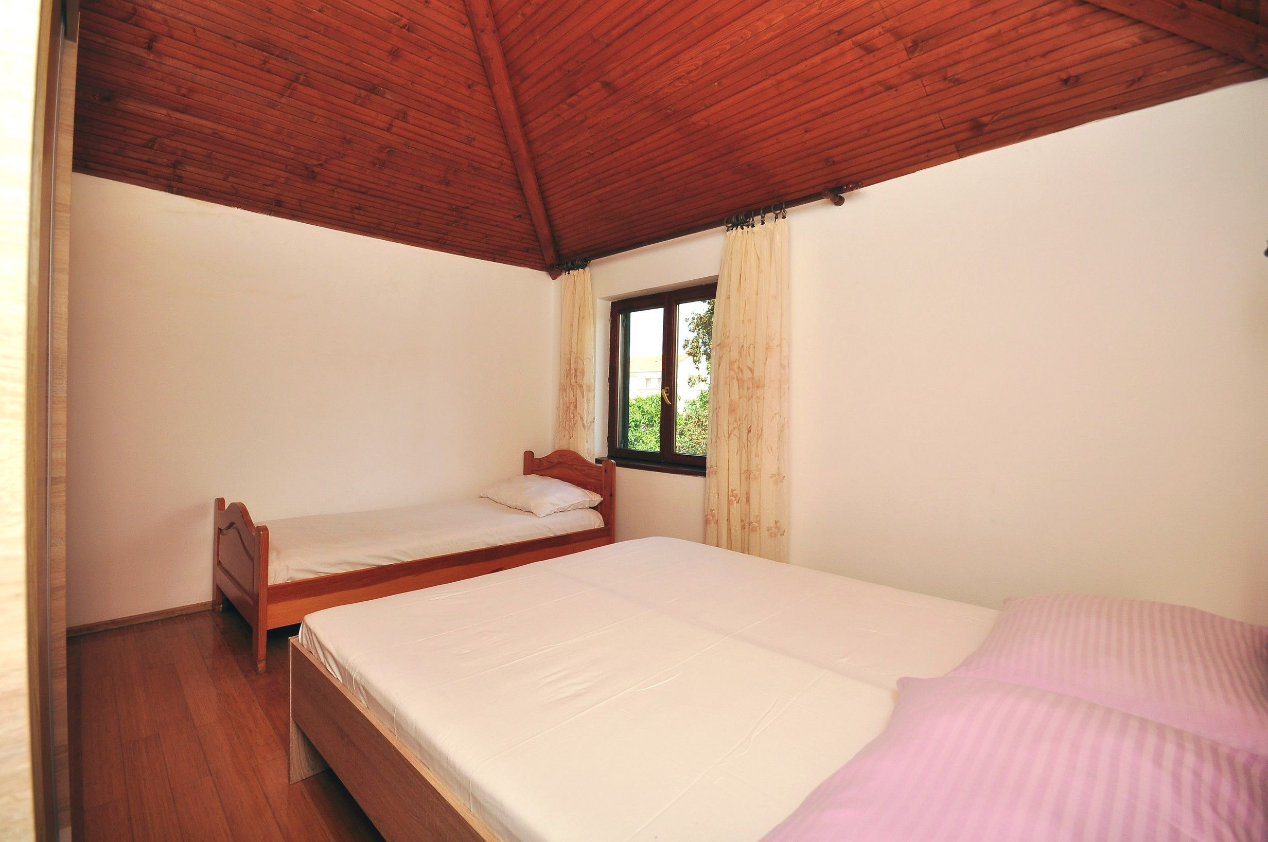 Une chambre avec deux lits simples, un plancher en bois, un plafond en bois et une fenêtre avec des rideaux.