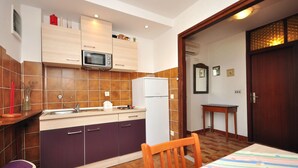 A compact kitchen with a microwave, refrigerator, and sink. There is a small wooden table and a chair. The kitchen has a tiled backsplash and wooden cabinets. - Apartments Marin (Novalja)