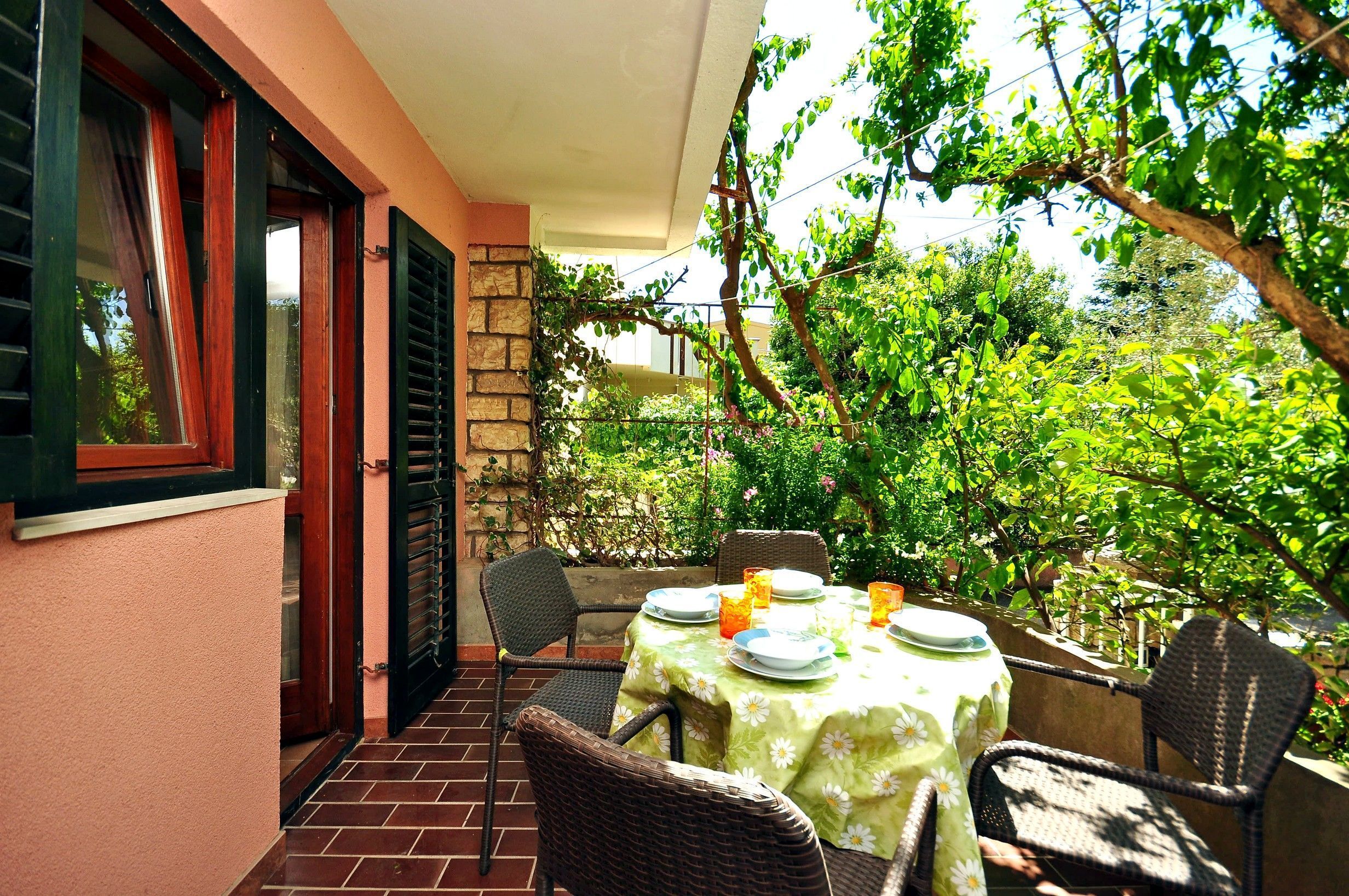 Un patio al aire libre con una mesa dispuesta para comer, rodeado de sillas de mimbre y un muro de piedra con vegetación.