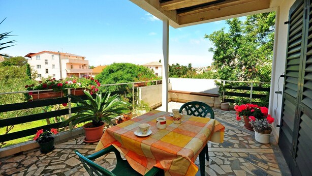 A covered outdoor patio with a table set for two, surrounded by potted plants and a view of the neighborhood. - Apartments Marin (Novalja)