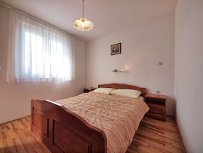 A bedroom with a wooden bed, a bedside table, a nightstand, a hanging light fixture, and a window with sheer curtains. - Apartments Vladimir (Vodice)