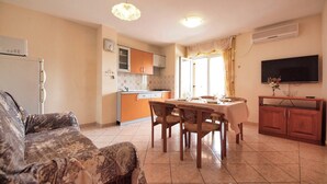 A living room with a sofa, a dining table with chairs, a kitchen area with cabinets, and a TV mounted on the wall. - Apartments Vladimir (Vodice)