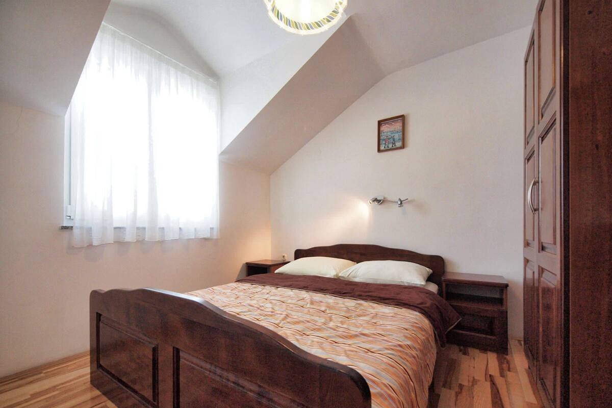 A bedroom with a wooden bed, a nightstand, a wall-mounted lamp, a window with sheer curtains, and a wooden wardrobe.