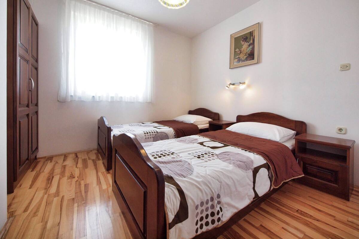 A bedroom with two wooden beds, a window with sheer curtains, a wall-mounted light, and a painting.