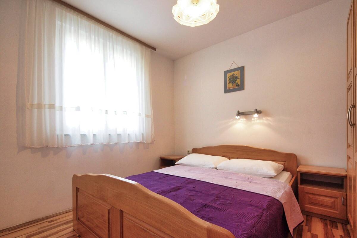 A bedroom with a wooden bed, a nightstand, a bedside table, a window with sheer curtains, a wall-mounted light fixture, and a framed picture.