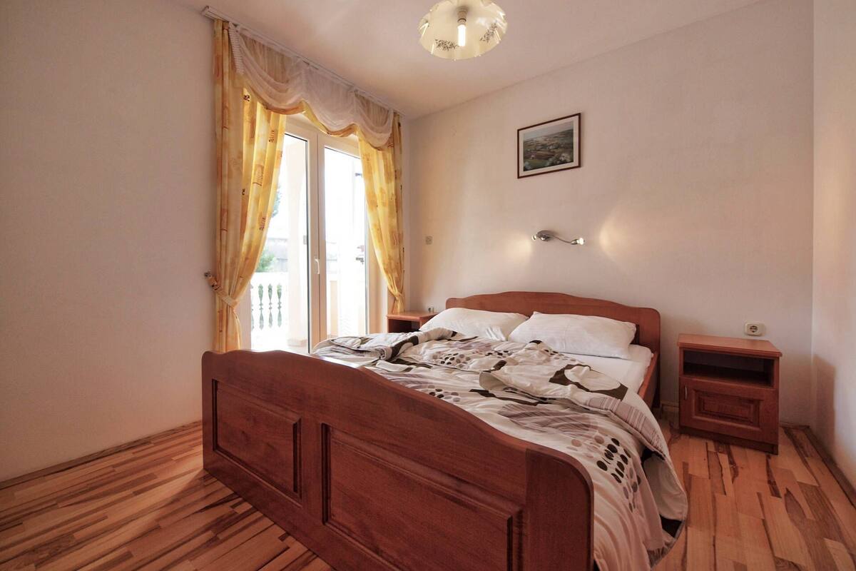 A bedroom with a wooden bed, a nightstand, a bedside table, a ceiling light, and a window with curtains.