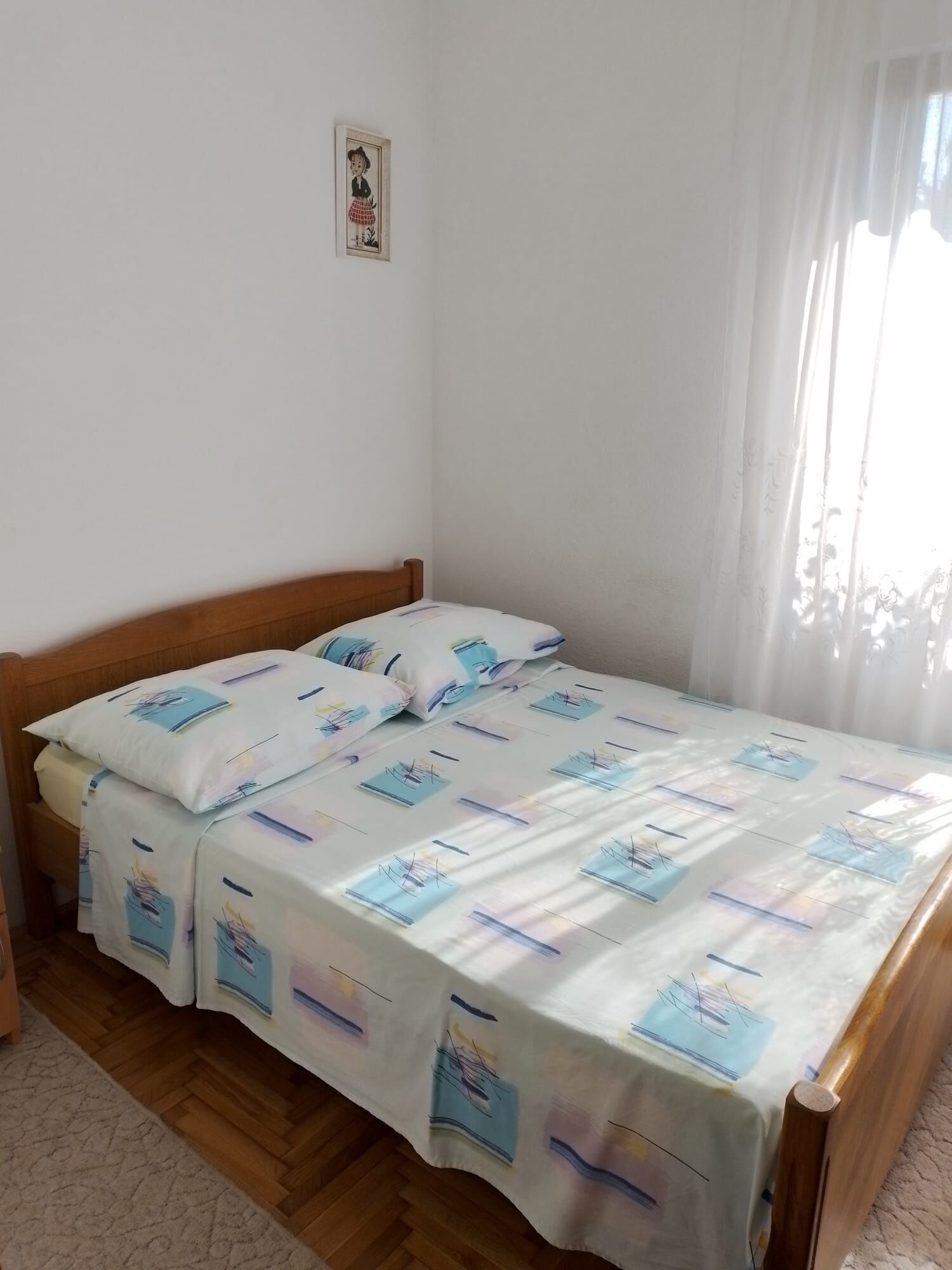 A single bed with a patterned bedspread, a wooden headboard, a small framed picture on the wall, and a window with sheer curtains.