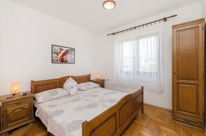 A bedroom with a wooden bed, a nightstand with a lamp, a wardrobe, and a window with sheer curtains. - Apartments Miljenka (Vodice)