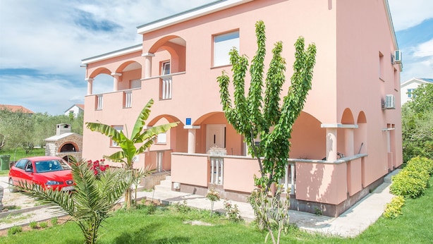 A pink two-story building with a stone oven, a red car parked in front, and a well-maintained garden. - Apartments Tatjana (Sukosan)