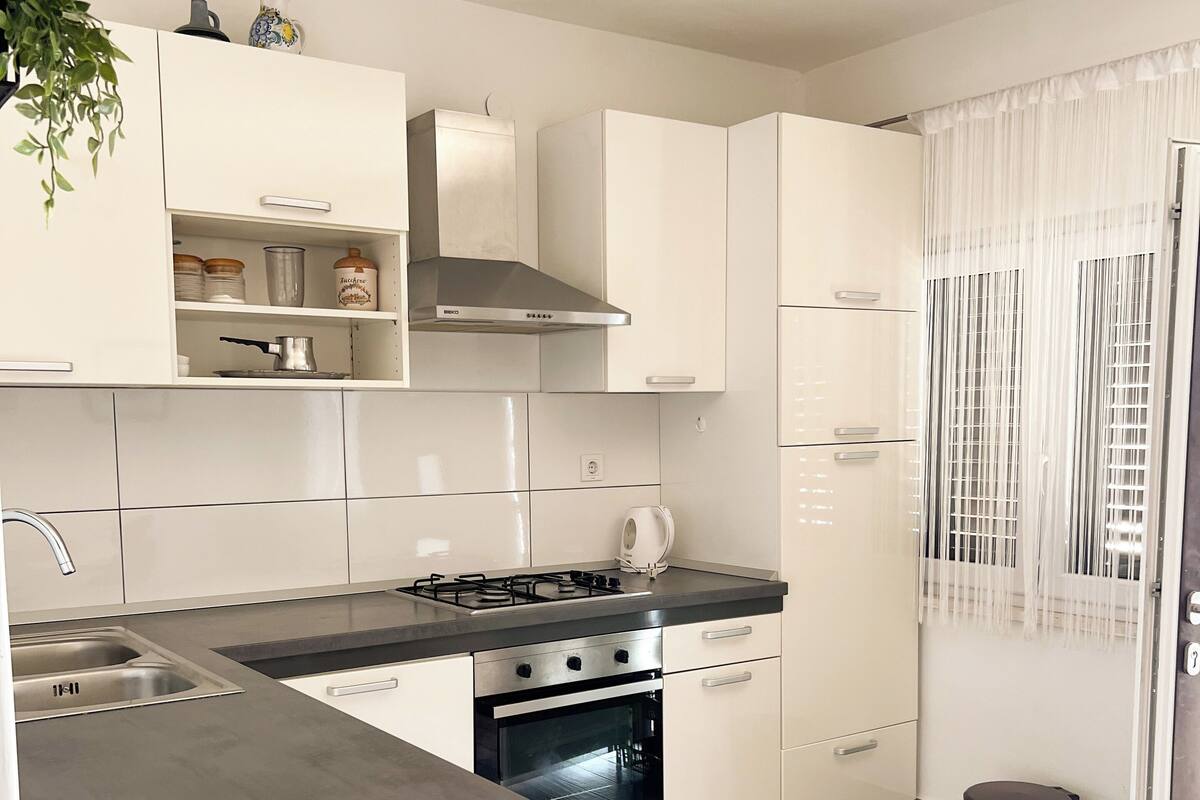 Une cuisine moderne avec des meubles blancs, une cuisinière en acier inoxydable et un four encastré. On trouve une poubelle et une plante en pot sur le plan de travail.