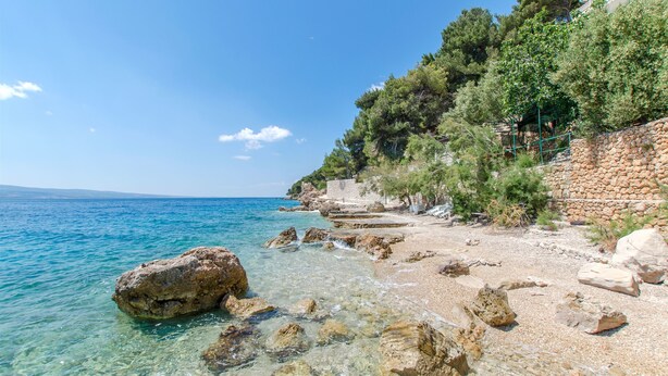 A coastal beach with clear waters, rocky shoreline, and a stone wall with vegetation. - Apartments Vedran (Stanici)