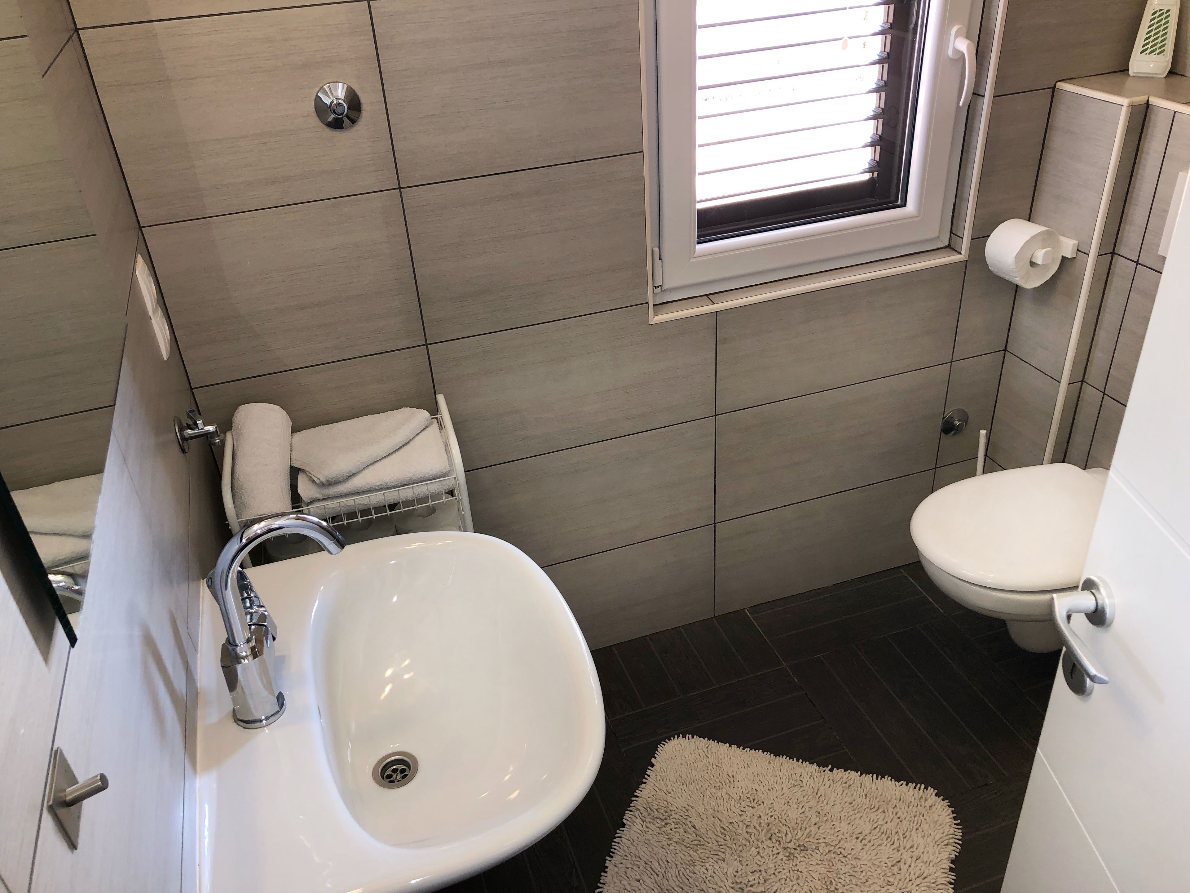 A modern bathroom with a white sink, toilet, and a small window with blinds.