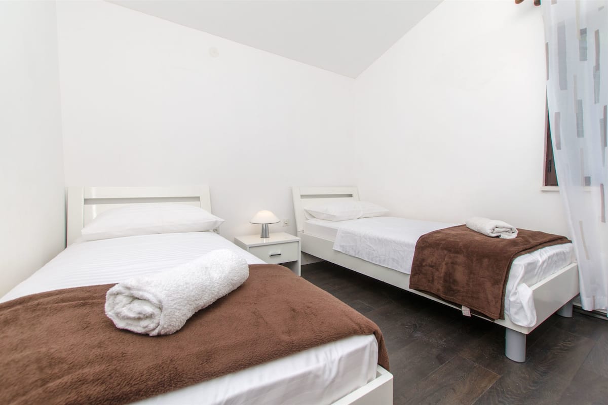 Une chambre d’hôtel avec deux lits, du linge de lit blanc, un plaid marron sur l’un des lits, une table de chevet avec une lampe et une fenêtre avec des rideaux.
